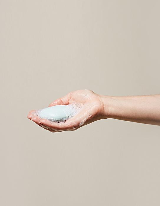 Single hand cupping the LATHER Avocado Mint Shampoo Bar, slightly sudsy as if just used against a beige background.
